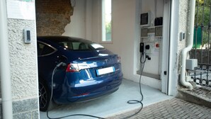 Un coche azul aparcado en una estación de carga de vehículos, con un muro de ladrillo y una ventana al fondo.