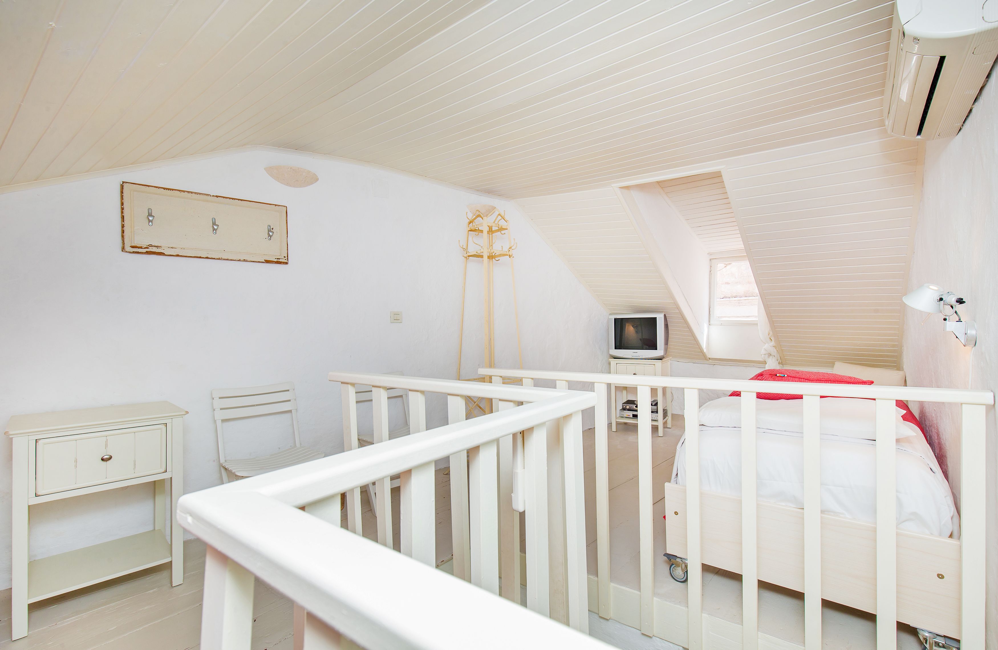Et soveværelse med skråvægg, en hvid seng, et sengebord, en stol, et fjernsyn og en lampe.