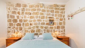 A bedroom with a stone wall, a bed with a white headboard, two bedside tables, and a small shelf with decorative items. - House Peti (Dubrovnik)