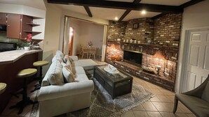 Ein Wohnzimmer mit Sofa, Couchtisch und Kamin. Im Hintergrund befindet sich eine Küchenzeile mit Schränken und einem Esstisch.