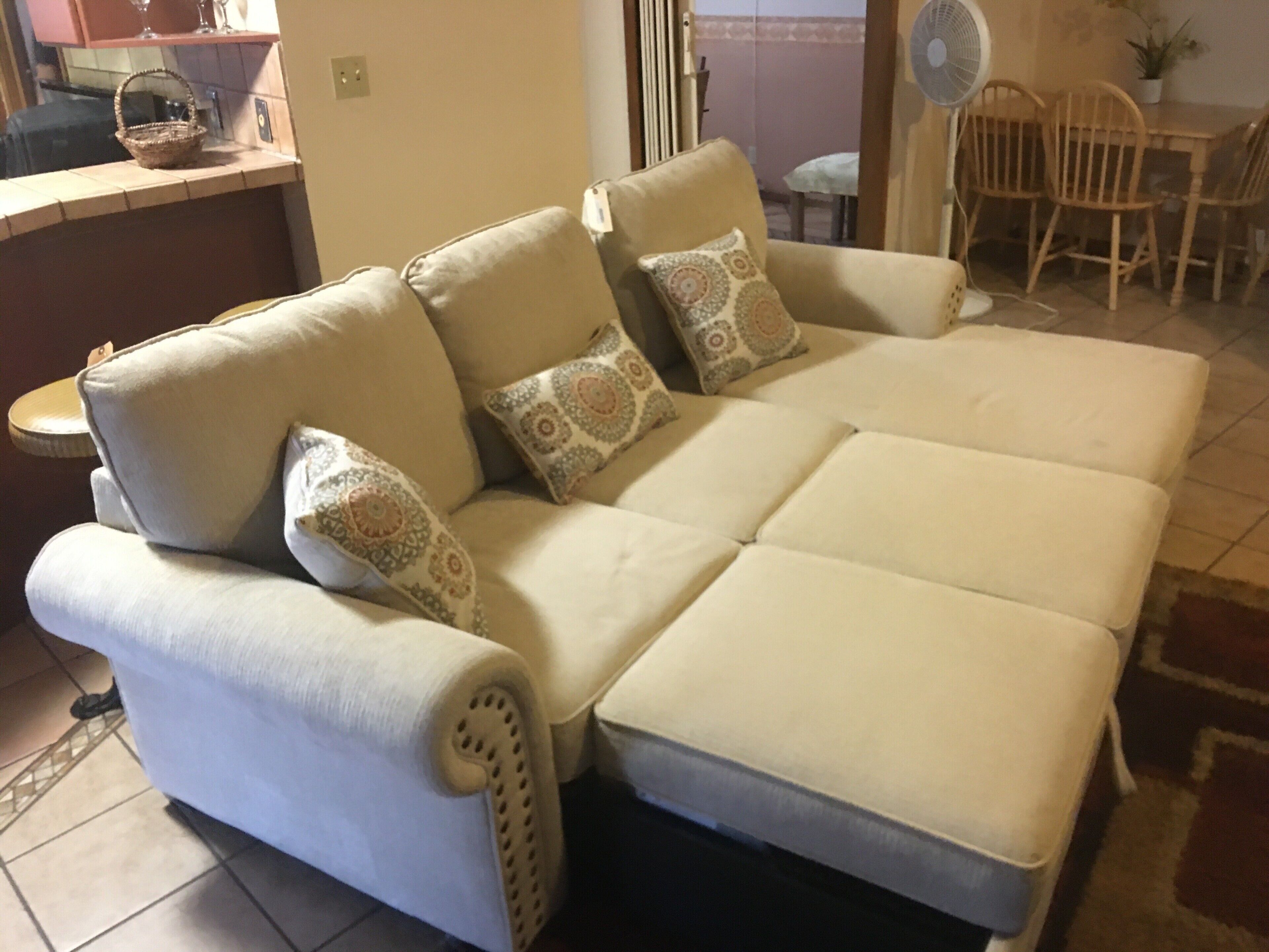 En beige hjørnesofa med mønstrede hynder, et lille sofabord og et spiseområde med træstole og en ventilator.