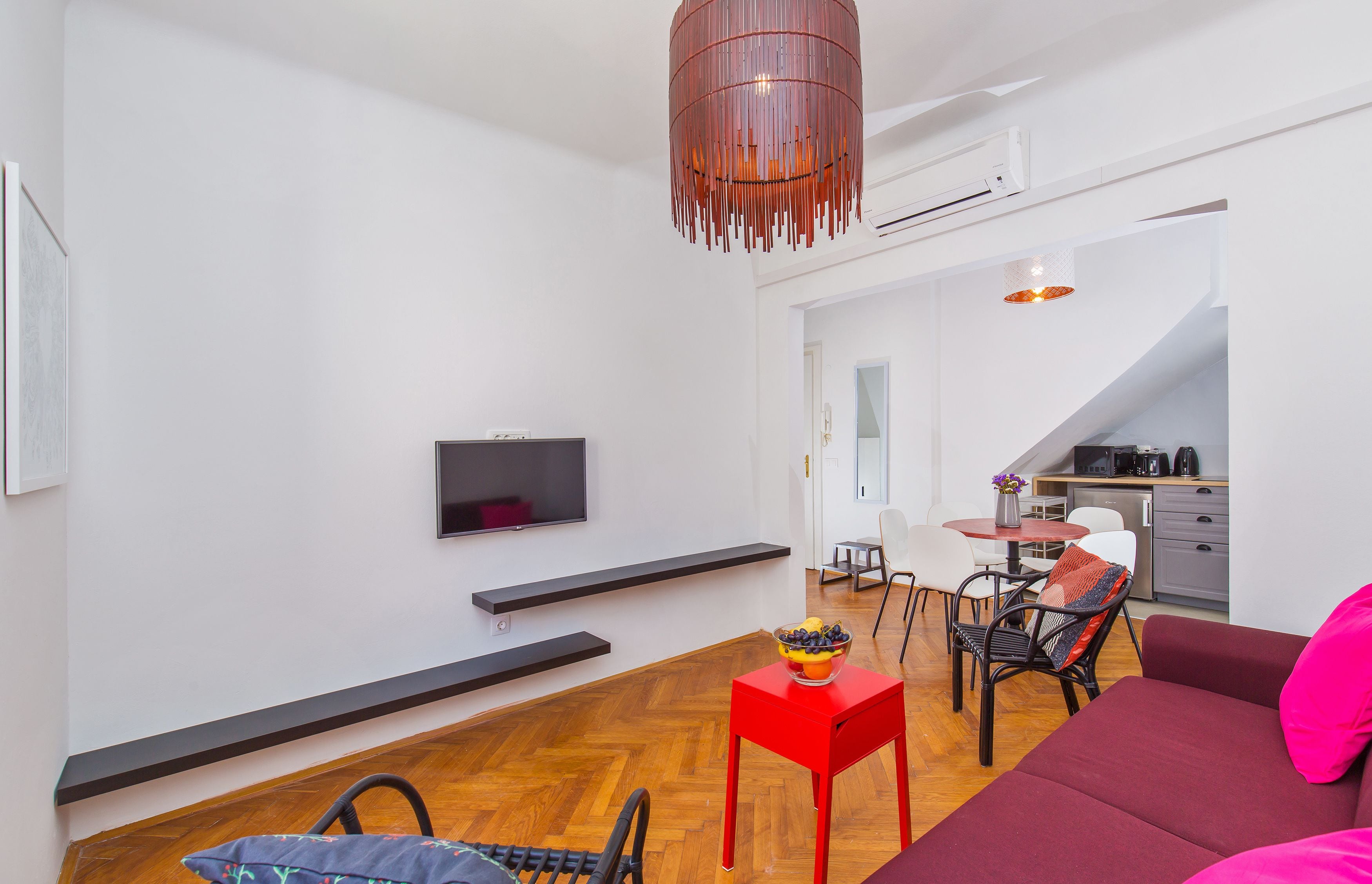 Un salon moderne avec un téléviseur à écran plat, une petite table d’appoint rouge sur laquelle se trouve un saladier de fruits, un canapé rouge et un coin repas avec une petite table et des chaises.