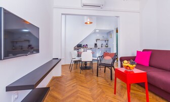 A modern living room with a flat-screen TV, a red table with a fruit bowl, a purple sofa, and a dining area with a white table and chairs.