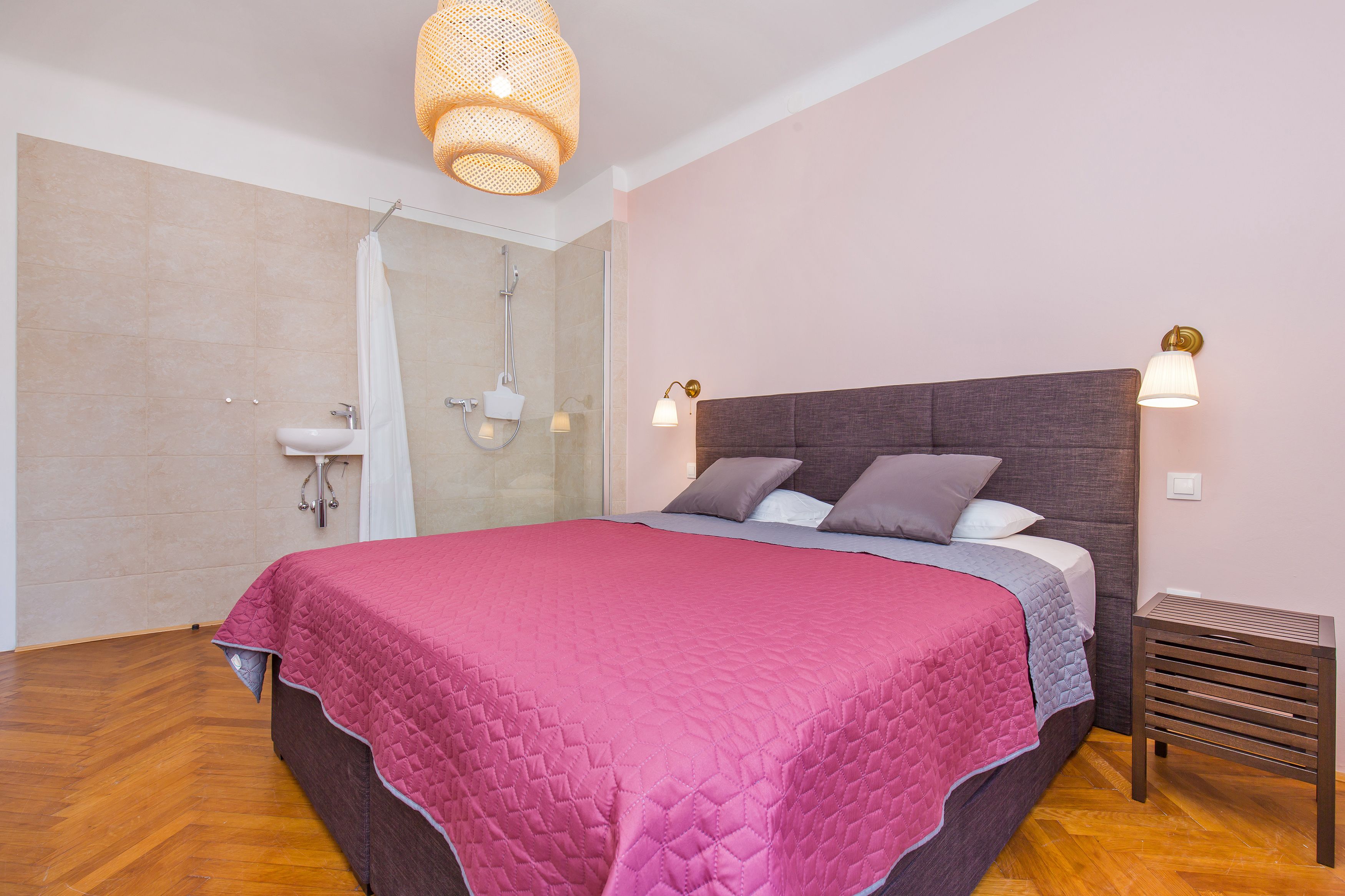 A bedroom with a large bed, a bedside table, a nightstand, a shower, and a bathroom sink.