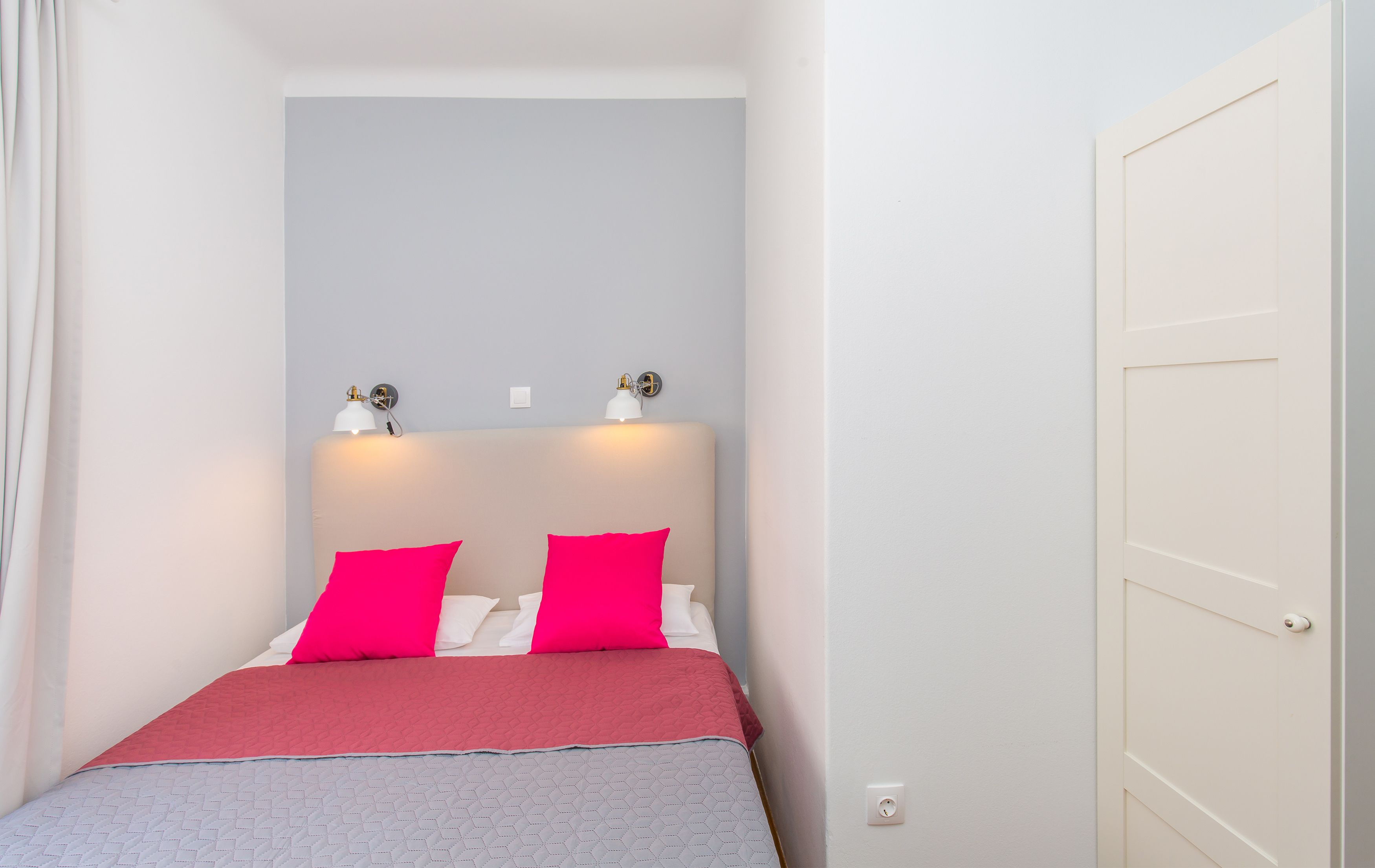 A small, neatly arranged bedroom with a bed, two bedside lamps, and a door.