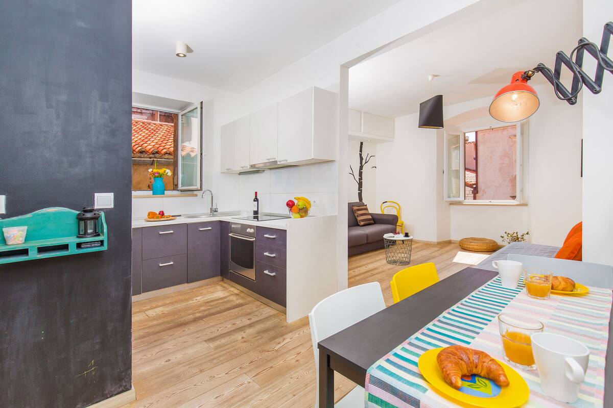 Una cocina moderna con armarios blancos, un comedor con mantel a rayas y vistas al exterior por una ventana.