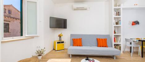 Una sala moderna con un sofá gris, cojines naranjas, un televisor de pantalla plana, un mueble amarillo pequeño y un otoman tejido.
