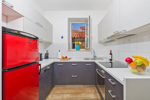 A compact kitchen with a red refrigerator, a small window with a view of a house, and a fruit bowl on the counter.