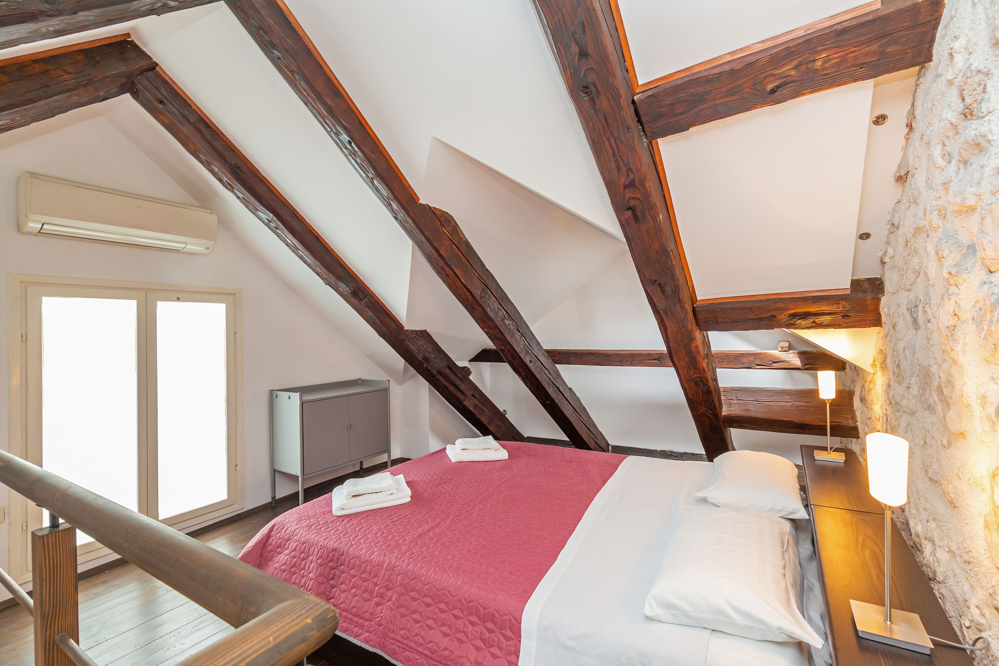 A bedroom with a sloped ceiling, wooden beams, a bed with a red quilt, white pillows, a bedside table with a lamp, and a window with blinds.