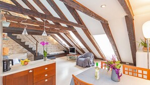 Un espace de vie de style « loft », avec des poutres en bois apparentes, une table à manger avec des chaises, un coin cuisine équipé d’une cuisinière et de placards, et un escalier menant à un niveau supérieur.