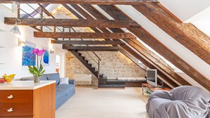 Une chambre mansardée avec des poutres apparentes, un mur en pierre, un escalier, un canapé, une télévision et un bureau avec des tiroirs.