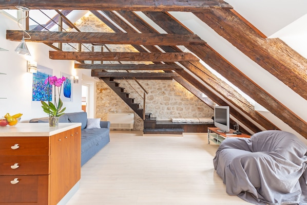 An attic room with exposed wooden beams, a stone wall, a staircase, a sofa, a TV, and a desk with drawers.