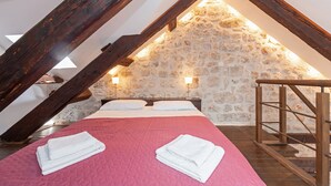 Une chambre confortable située dans les combles, avec un mur en pierre, des poutres apparentes, un lit recouvert d’une couette rose et deux lampes de chevet.