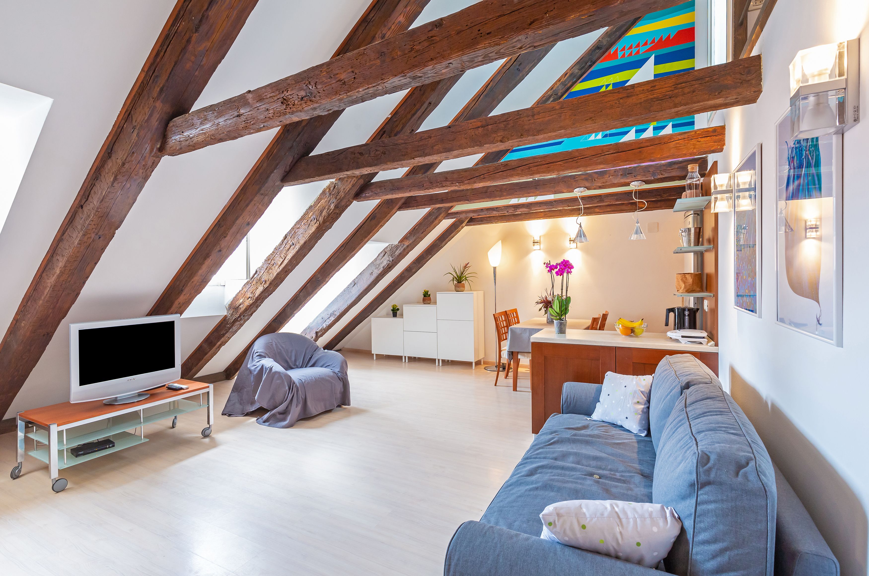 Una sala de estar estilo loft con vigas de madera a la vista, un televisor de pantalla plana sobre un soporte móvil, un sofá gris y una zona de comedor con una pequeña mesa y sillas.
