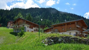 A wooden chalet-style building with balconies, surrounded by a grassy area and a stone wall, set against a backdrop of a forested mountain. - Proche du Centre! Appart sympa, Piscine + Sauna (Valfréjus)