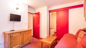 A compact room with a TV mounted on the wall, a wooden cabinet, a small table, and a red sofa. - Proche du Centre! Appart sympa, Piscine + Sauna (Valfréjus)