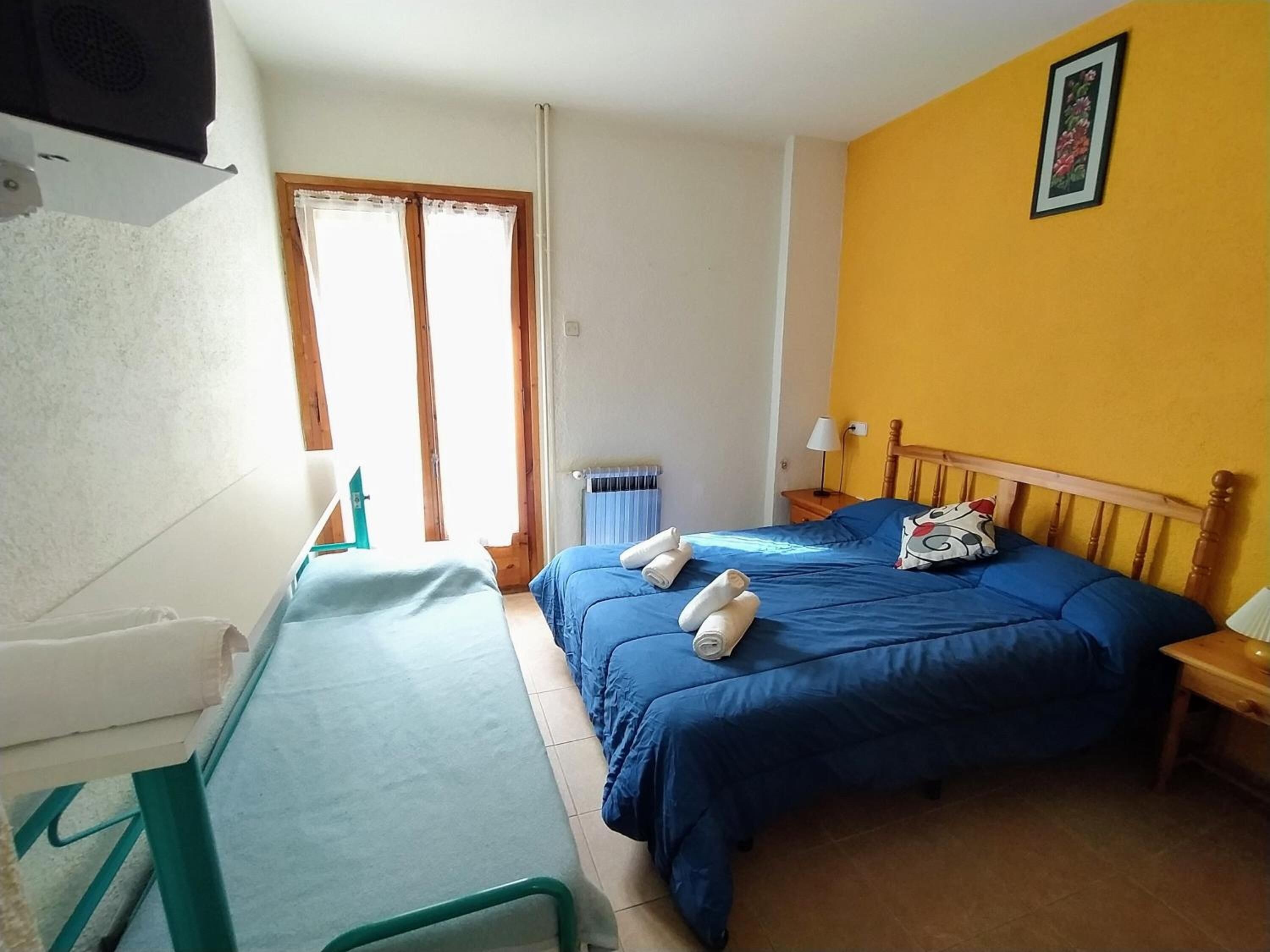 A bedroom with two beds, a window with curtains, a radiator, and a bedside table with a lamp.