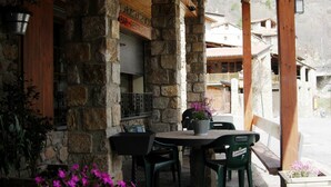 A stone building with a wooden roof, a table with chairs, and a potted plant with pink flowers. - Hostal Pas de la Pera (Lles)