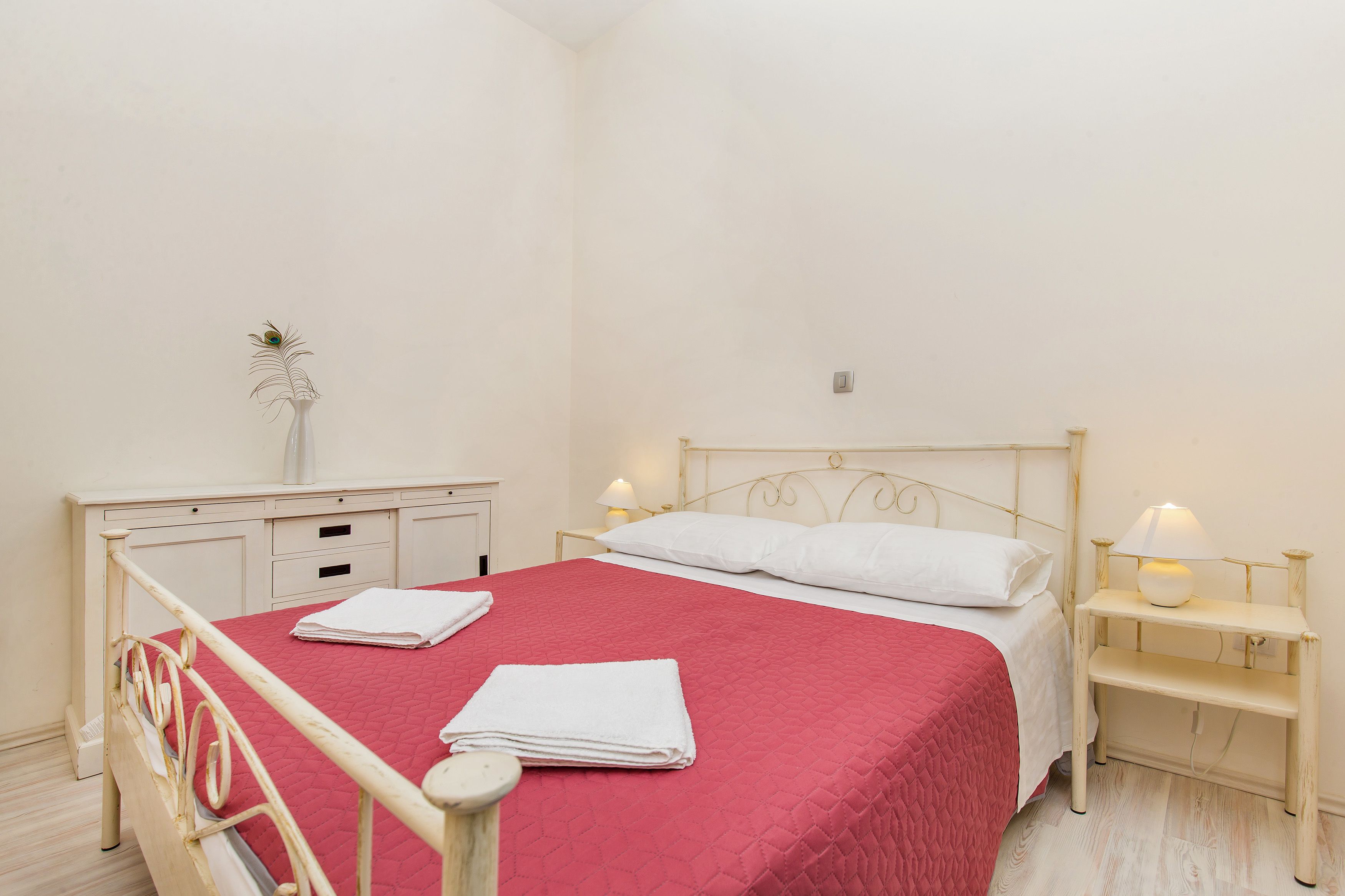 A neatly made bed with a red quilt, white pillows, and folded towels. There is a bedside table with a lamp, a small white vase with a plant, and a wooden nightstand with a lamp.