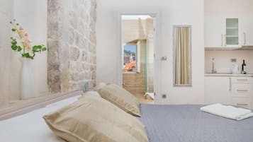 A bedroom with a bed, pillows, a vase with flowers, a stone wall, a doorway to a balcony, and a kitchen area with cabinets and a wine bottle.
