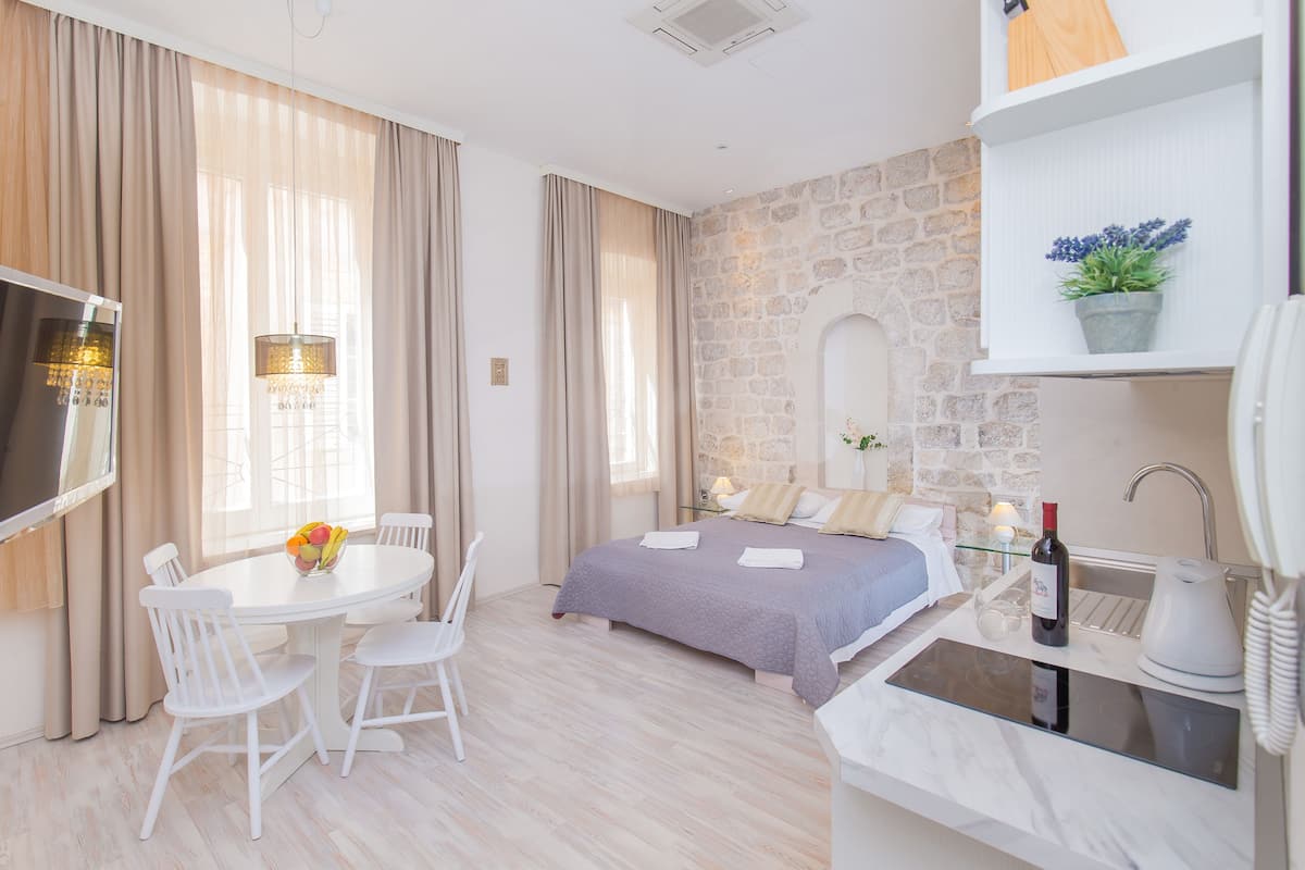 A modern hotel room with a stone accent wall, a bed with a purple bedspread, a dining area with a white table and chairs, and a kitchenette with a wine bottle.