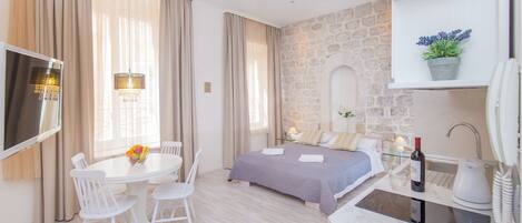Una habitación de hotel moderna con una pared de acento de piedra, una cama con colcha morada, un área de comedor con una mesa y sillas blancas, y una kitchenette con una botella de vino.