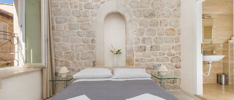 A bedroom with a stone wall, a bed with white linens, a glass table with a lamp, and a bathroom visible through an open door.