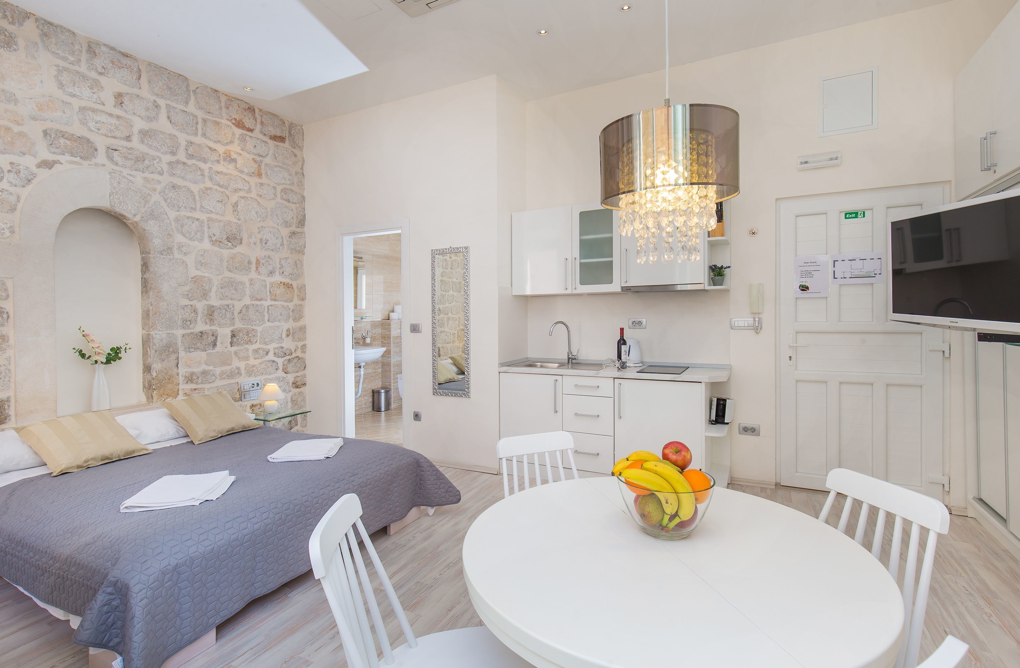 Un dormitorio moderno con pared de piedra, cama con colcha gris, mesa de comedor redonda con fruta y cocina con armarios blancos y televisor.