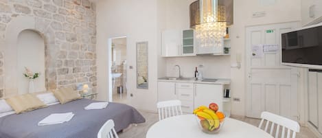 Un dormitorio moderno con pared de piedra, cama con colcha gris, mesa de comedor redonda con fruta y cocina con armarios blancos y televisor.
