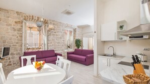 A modern living room with a stone wall, a purple sofa, a white dining table with fruit, and a kitchen with a sink and utensils.