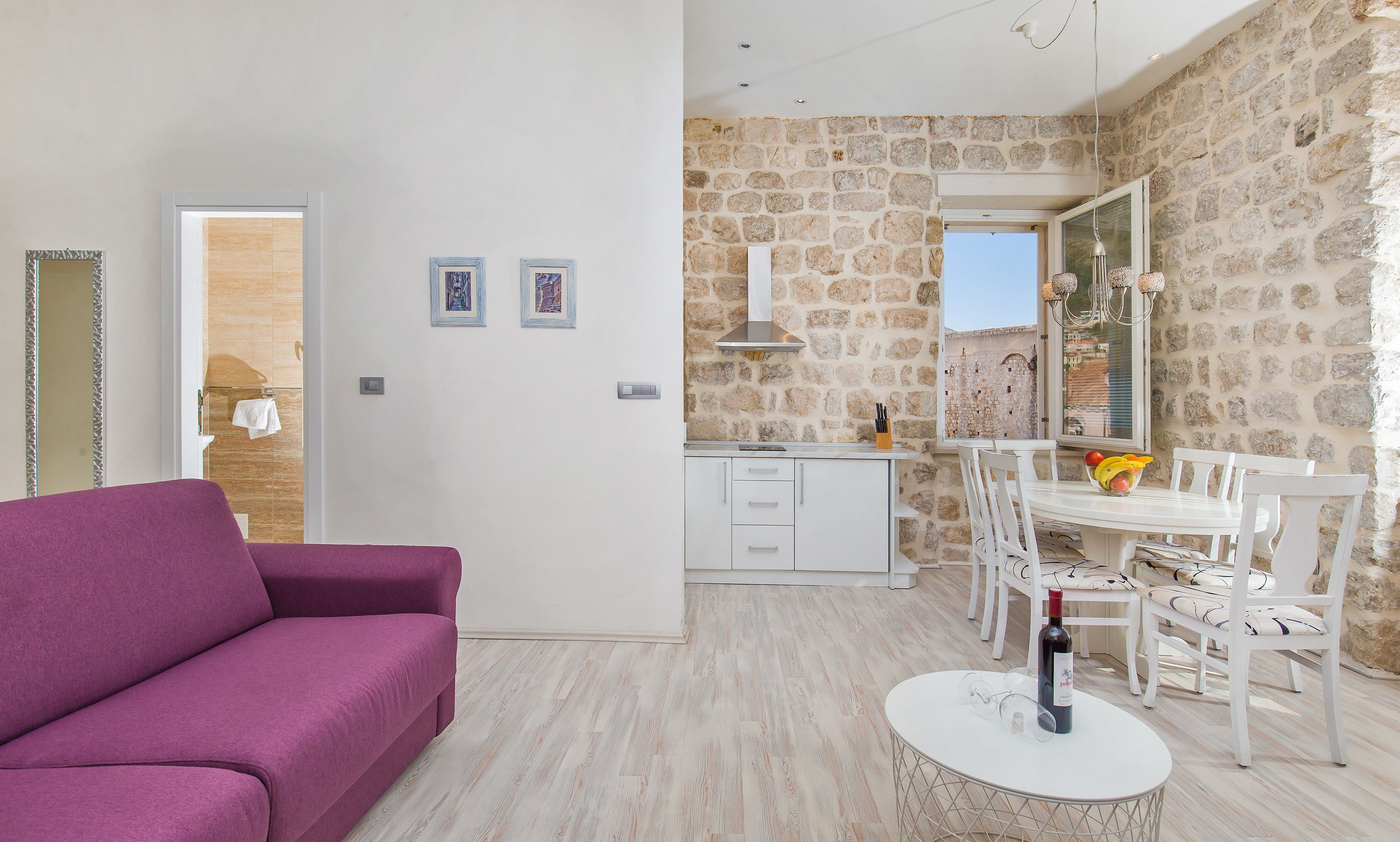 A modern living room with a purple sofa, a dining area with a white table and chairs, and a kitchen with stone walls and a window.