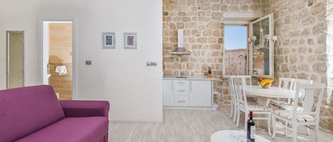 Una sala de estar moderna con un sofá púrpura, una zona de comedor con una mesa y sillas blancas, y una cocina con paredes de piedra y una ventana.