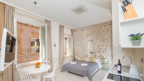 A modern room with a stone wall, a bed with a gray bedspread, a small kitchen area with a sink and a wine bottle, and a dining area with a table and chairs.