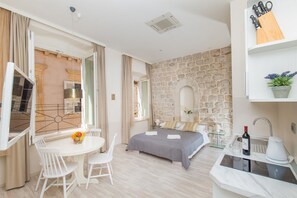 A modern room with a stone wall, a bed with a gray bedspread, a small kitchen area with a sink and a wine bottle, and a dining area with a table and chairs. - Apartments Lausion (Dubrovnik)