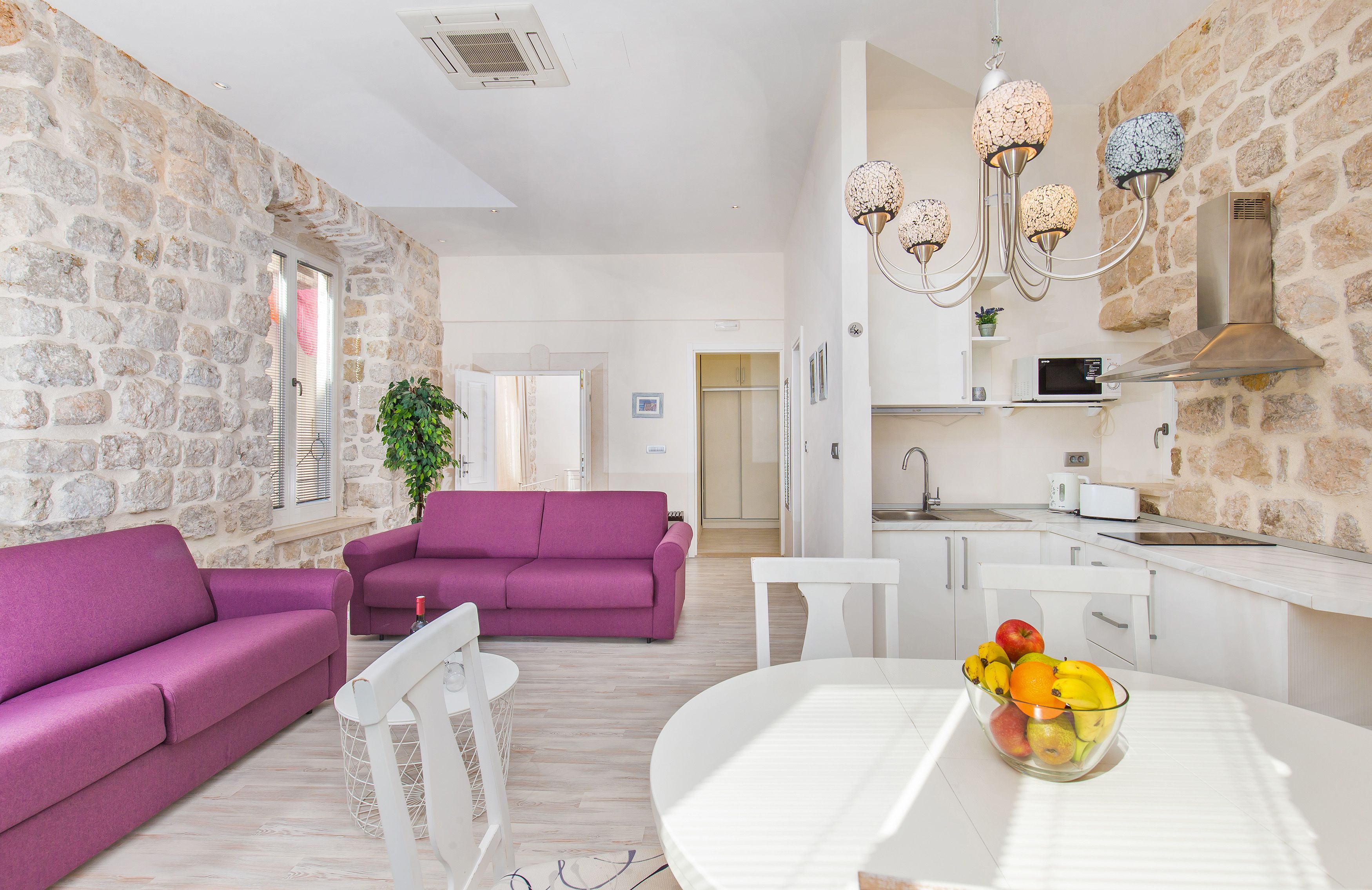A modern living room with a purple sofa, a dining table with a fruit bowl, and a kitchen area with a stone wall and white cabinets.