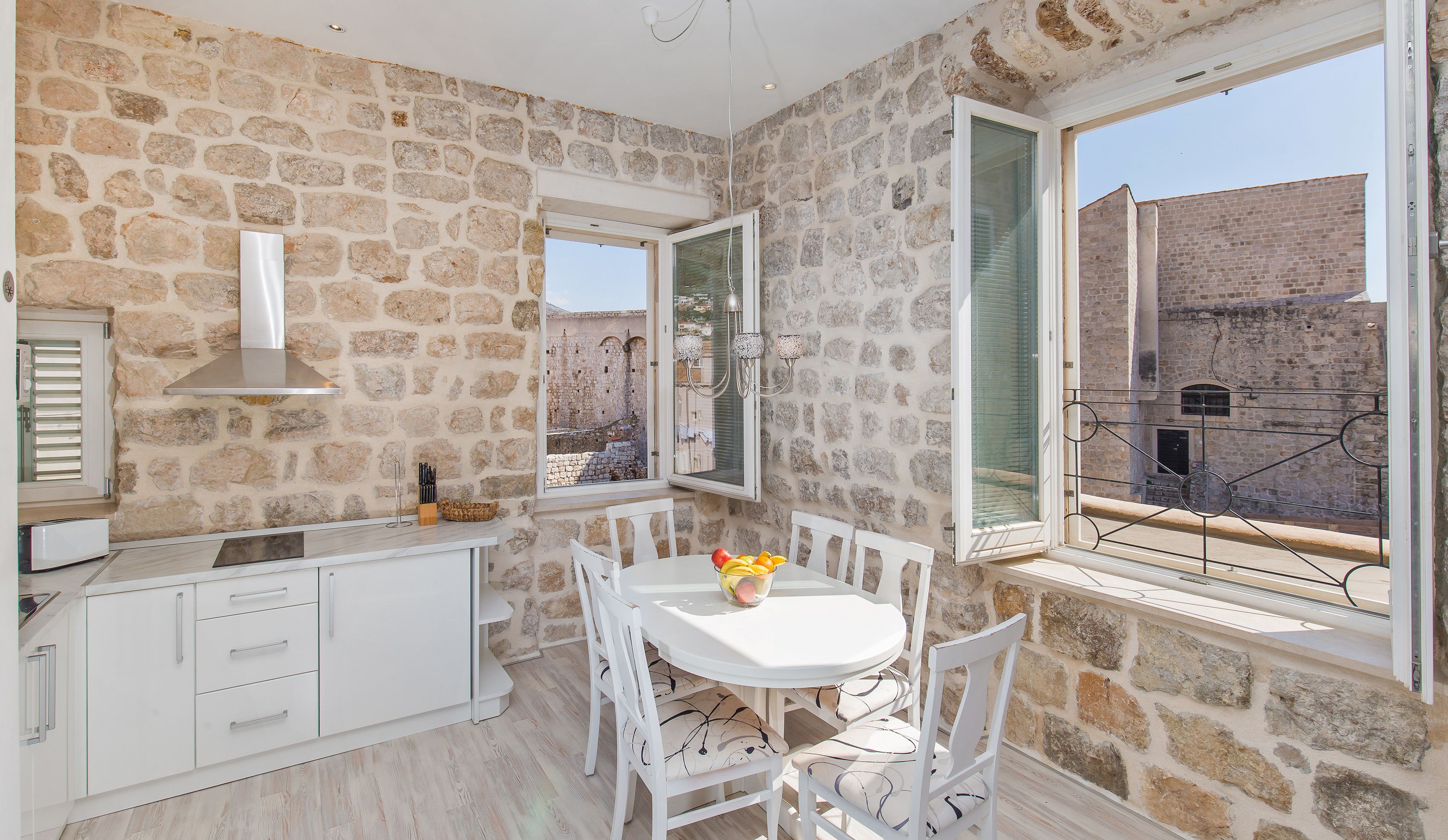 Une cuisine aux murs de pierre, dotée de meubles blancs, une table à manger avec des chaises, et une vue sur un bâtiment historique à l’extérieur.