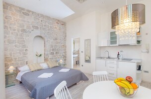 Quarto com parede de pedra, uma cama, mesa de cabeceira com abajur, banheiro visível através de um arco, área de cozinha com pia e armários, e uma fruteira sobre uma mesa.