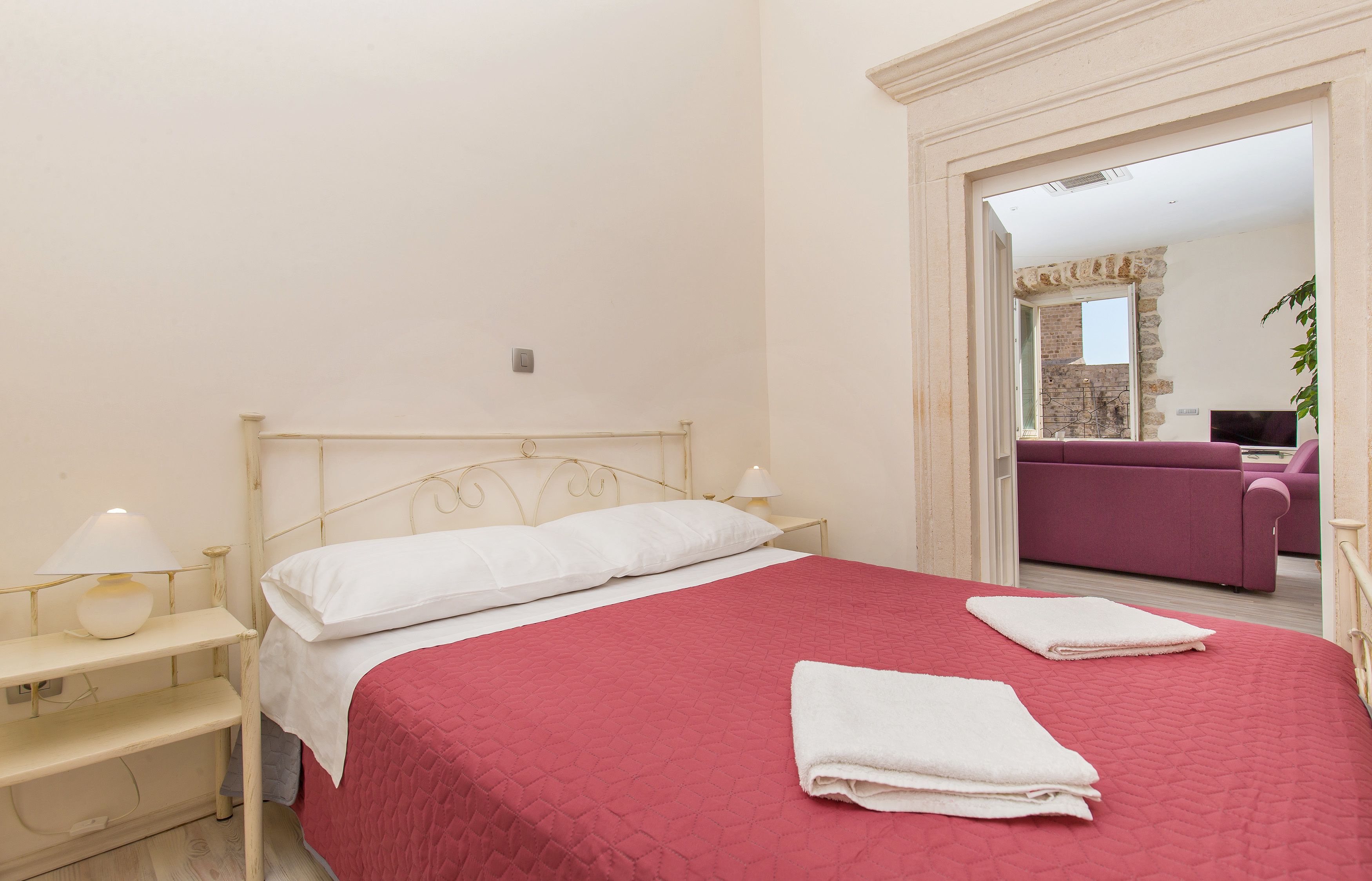 A neatly made bed with a red quilt, white pillows, and two folded white towels. There is a nightstand with a lamp and a shelf below the bed. A doorway leads to a room with a purple sofa and a television.