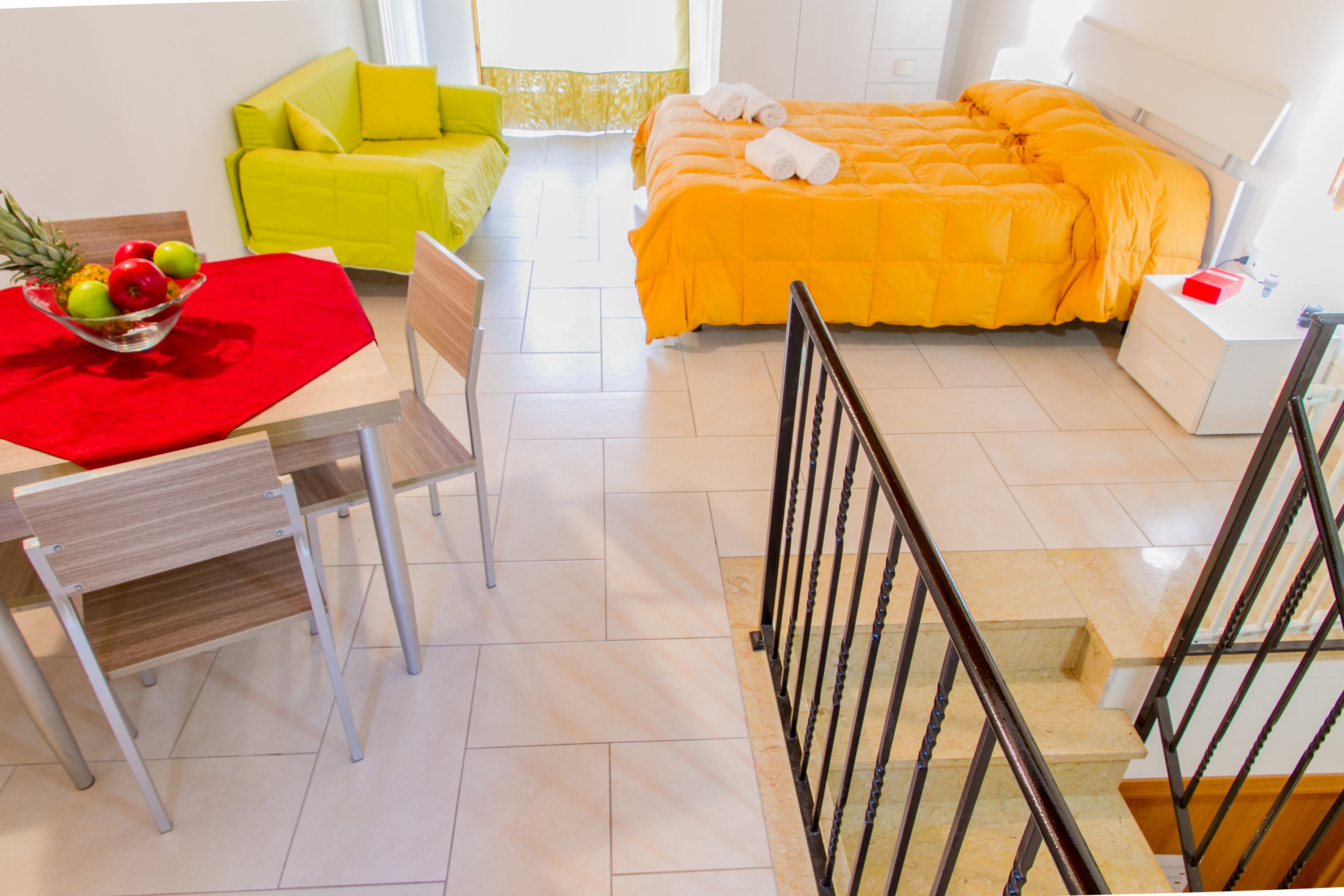 Quarto de hotel com uma cama, uma mesa de jantar com toalha de mesa vermelha, um sofá verde e uma escada.