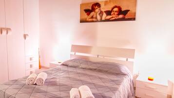 A neatly made bed with a quilted grey bedspread and two rolled towels on it. There is a white nightstand with a lamp and a small orange object on it. A painting of cherubs is on the wall above the bed.