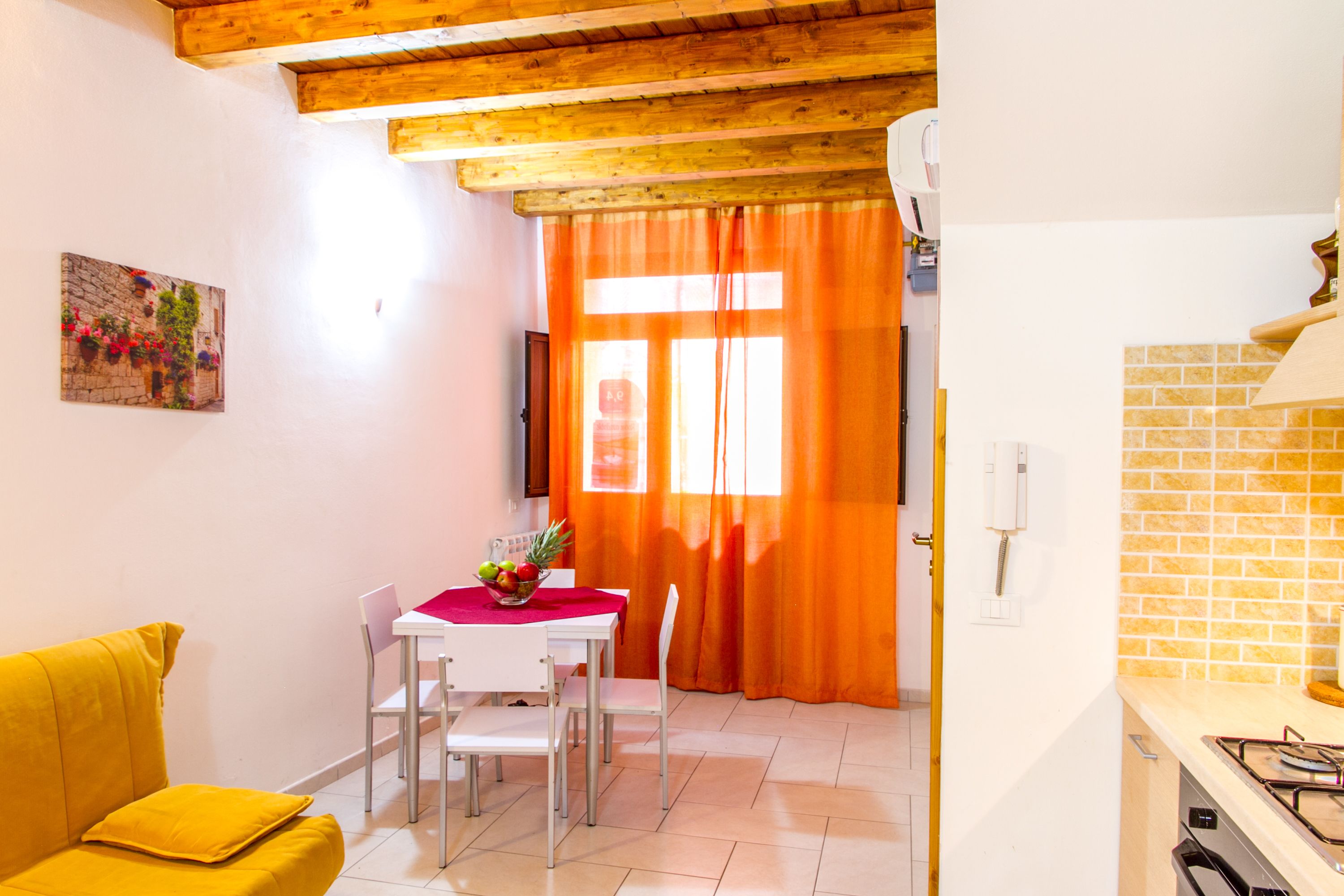 A dining area with a white table, four chairs, and a red tablecloth. There is a yellow sofa with a cushion, a painting on the wall, and a kitchen area with a tiled backsplash and a stove.