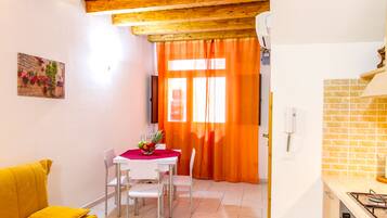 Un comedor con una mesa blanca, cuatro sillas y un mantel rojo. Hay un sofá amarillo con un cojín, un cuadro en la pared y una zona de cocina con salpicadero de azulejos y una estufa.