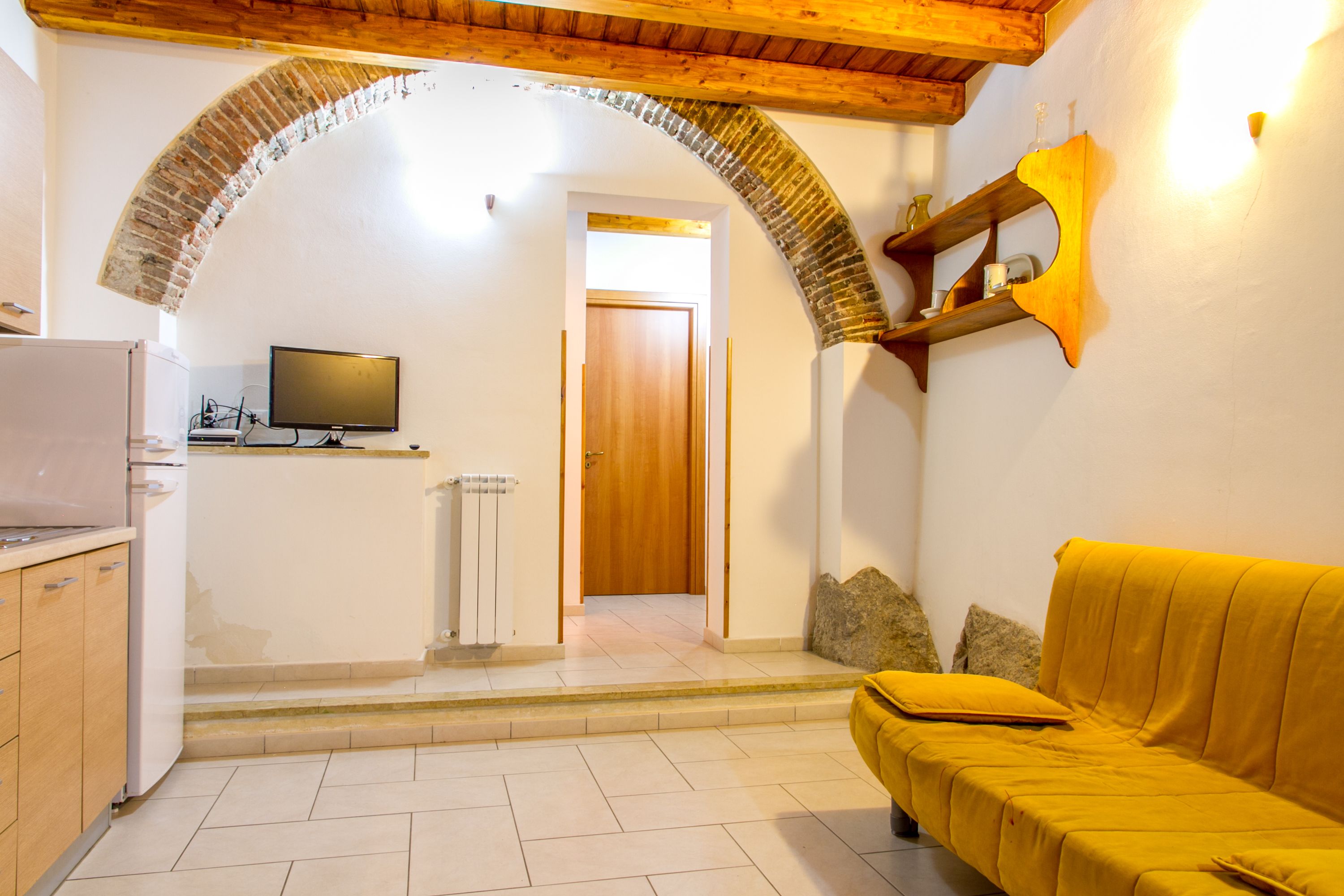 Una habitación con techo de madera, un arco de ladrillo, un sofá amarillo, un televisor y una pequeña cocina.