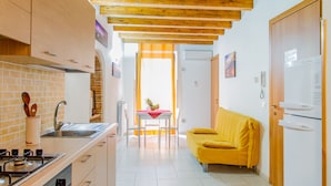Una cocina compacta con estufa, lavamanos y vigas de madera en el techo. Un sofá amarillo y una mesa de comedor con sillas se encuentran en la zona de estar contigua.