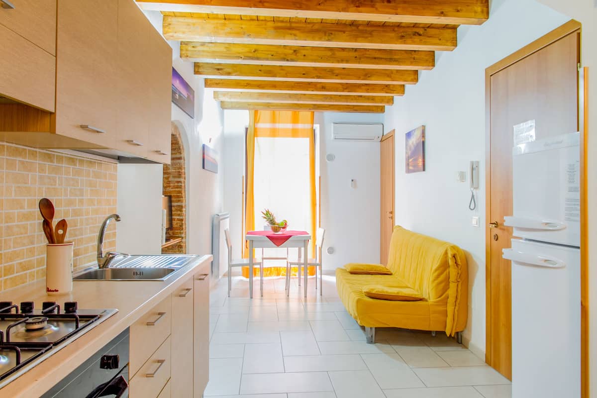 Una cocina compacta con estufa, lavamanos y vigas de madera en el techo. Un sofá amarillo y una mesa de comedor con sillas se encuentran en la zona de estar contigua.