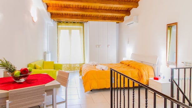 Una habitación luminosa con techo de madera, una cama con colcha naranja, una silla amarilla, una mesa con mantel rojo y un cuenco de fruta.