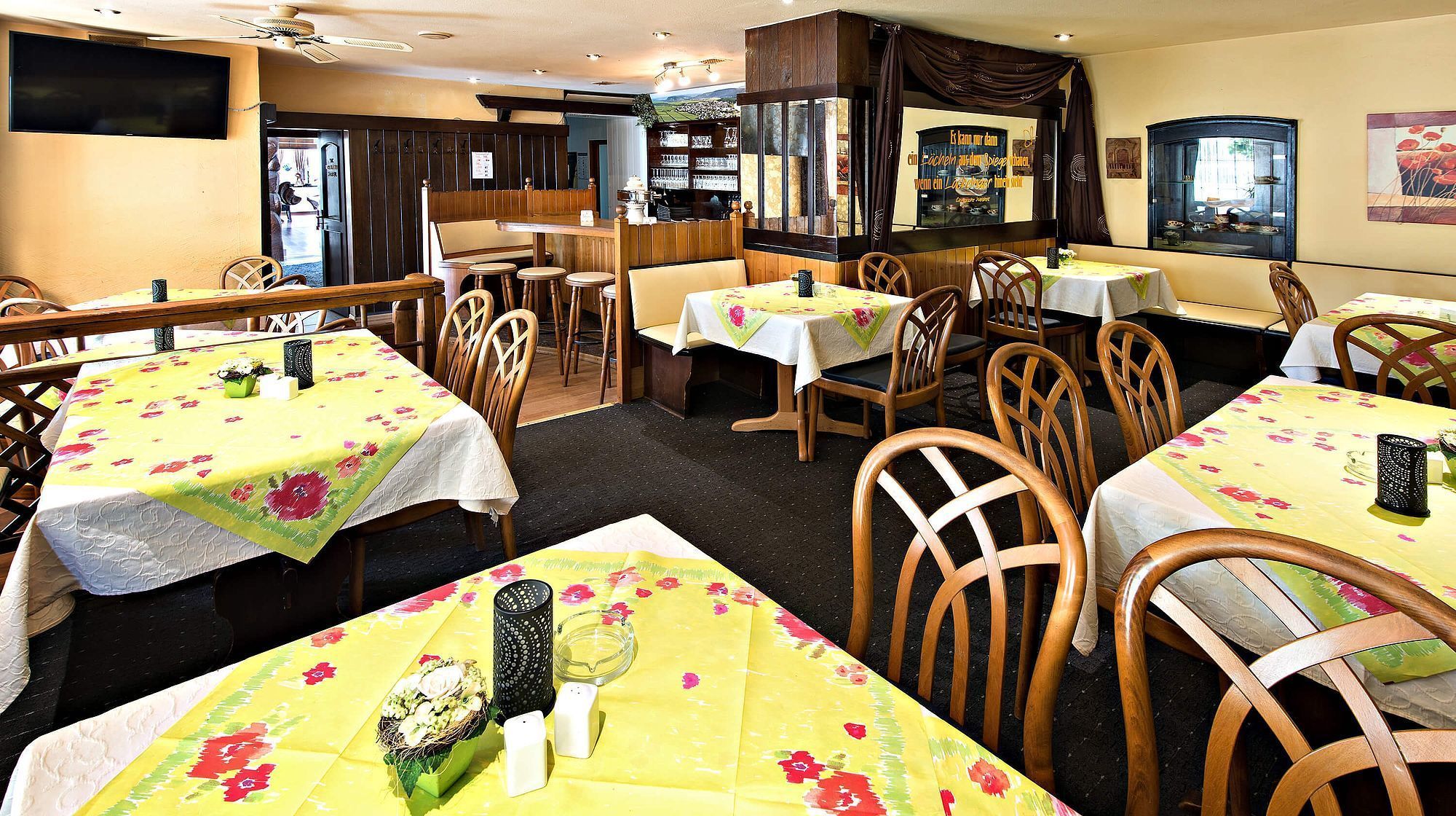Un ristorante con diversi tavoli rivestiti da tovaglie fiorate gialle, sedie di legno e un'area bar sullo sfondo.