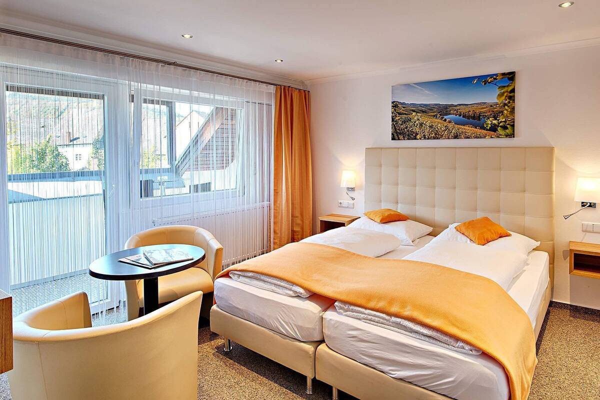 Een hotelkamer met een bed, een stoel, een klein tafeltje, een raam met gordijnen, en een schilderij aan de muur.