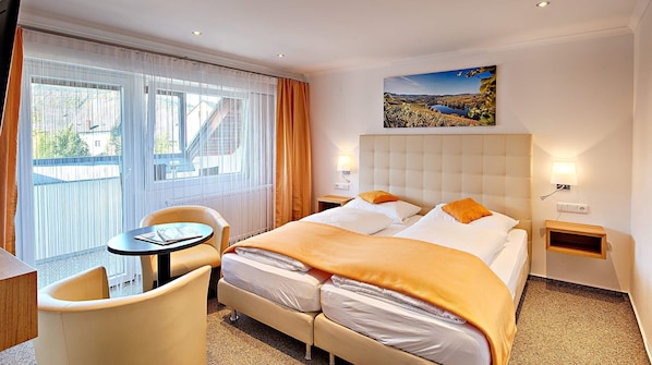 A hotel room with a bed, a chair, a small table, a window with curtains, and a painting on the wall.
