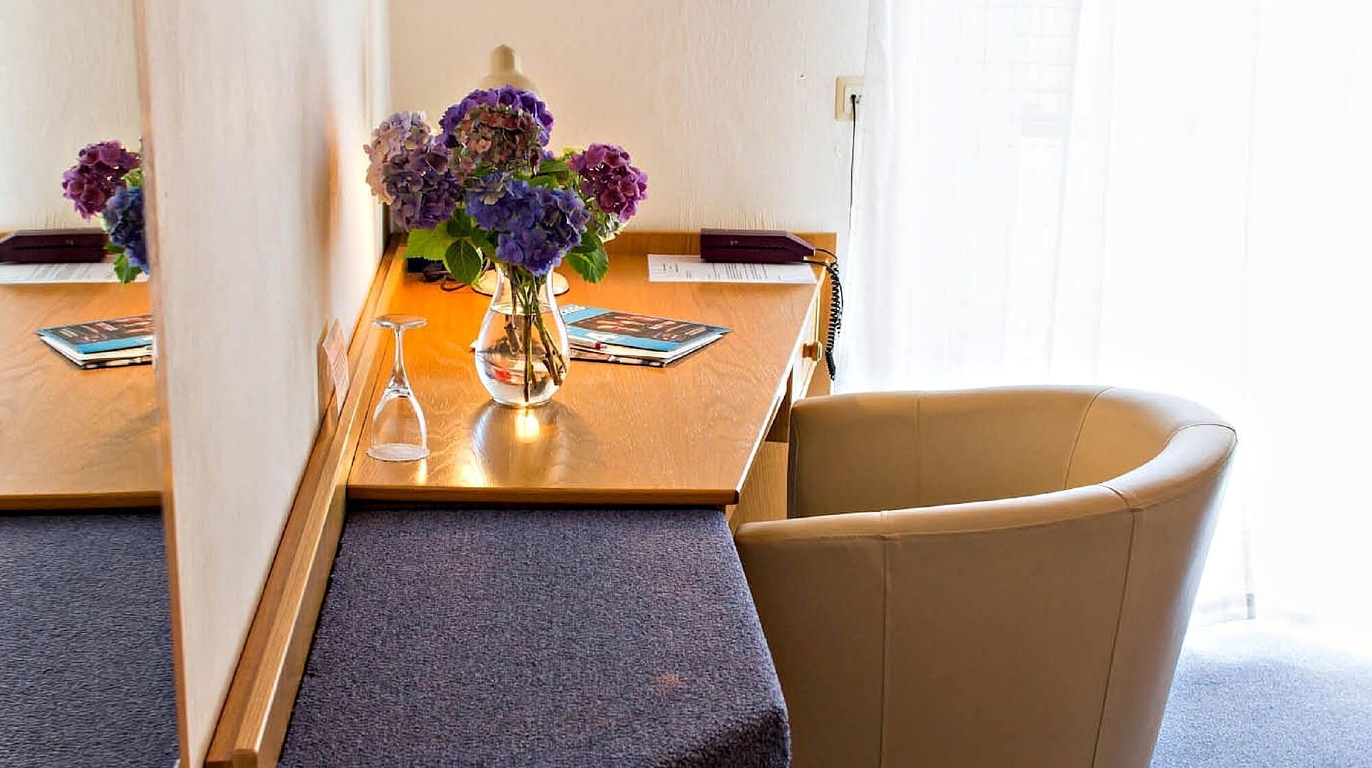 Quarto de hotel com uma mesa de madeira, um vaso com flores roxas, um copo e uma cadeira.